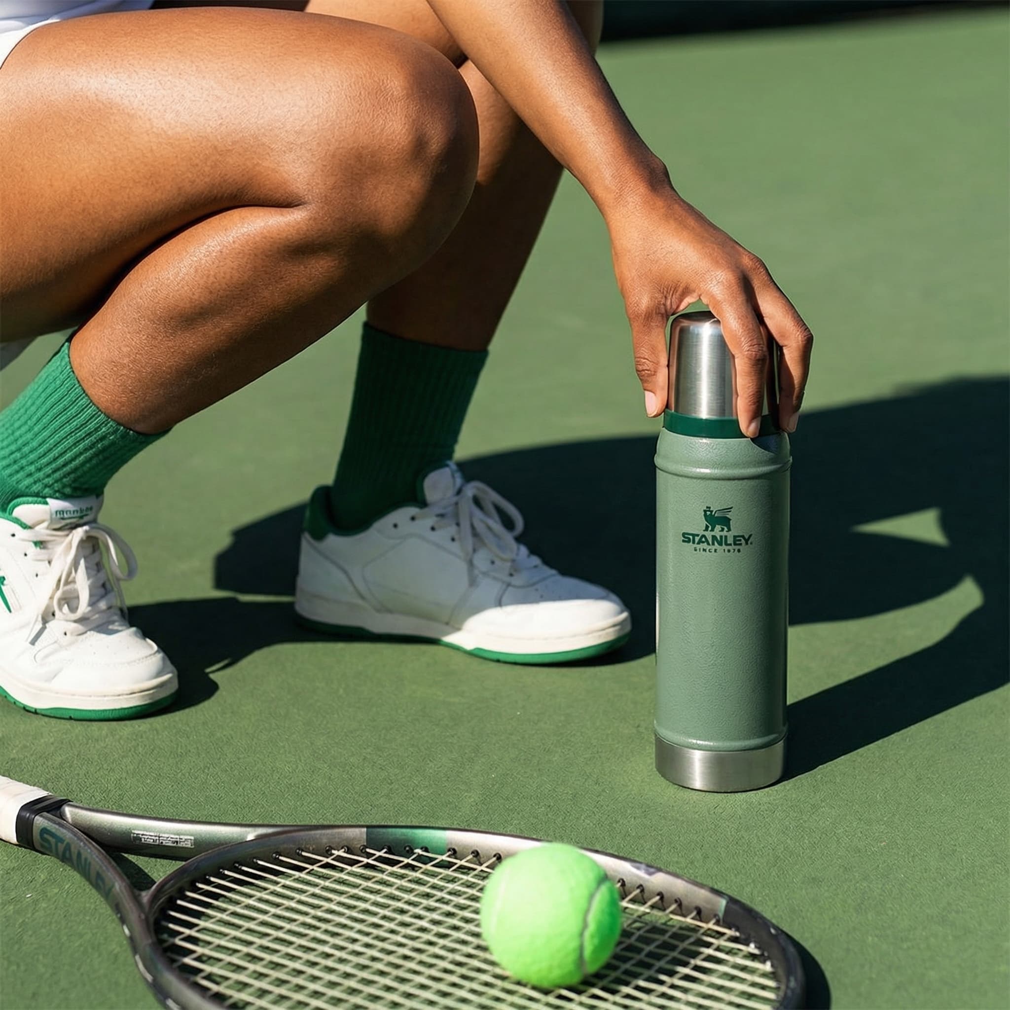 The image is a playful, sport-inflected lifestyle product shot that blends leisure, movement, and refreshment.
A person crouches on a tennis court, reaching down to place or pick up a slim canned drink positioned upright on the ground. The framing is tight on the lower body and hand, emphasizing action and context rather than identity. A tennis racket and bright green ball sit in the foreground, anchoring the scene clearly in an athletic setting.
Styling details—green ribbed socks, white sneakers with green accents—echo the color of the court and the can’s label, creating a cohesive, intentional palette. The can feels integrated into the moment, like a natural pause during play rather than a staged prop.
Lighting is strong and direct, likely midday sun, producing crisp shadows and saturated color. The texture of skin, court surface, and equipment is clearly visible, reinforcing an outdoorsy, tactile feel.
Overall, the image feels fun, casual, and contemporary, positioning the product as part of an active, social lifestyle—refreshment woven into sport and downtime rather than presented as a standalone hero object.