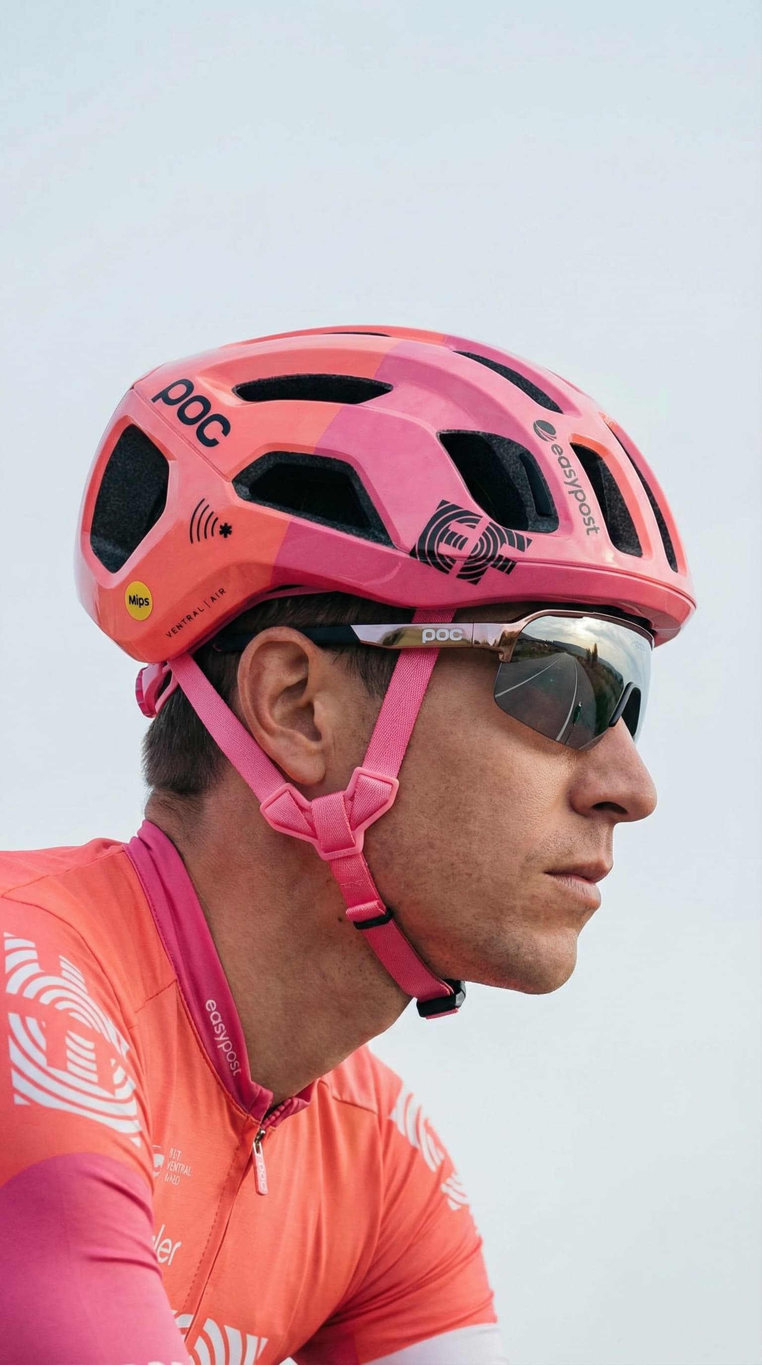 The image is a clean, performance-focused sports portrait with a sharp, modern edge.
A cyclist is shown in side profile, tightly framed from the shoulders up. The bright coral helmet and matching kit dominate the composition, immediately signaling speed, visibility, and technical design. The helmet’s vents, decals, and structured form emphasize safety and aerodynamics rather than lifestyle styling.
Mirrored wraparound sunglasses reflect the surrounding environment, adding a dynamic, almost futuristic feel while obscuring the eyes. This shifts the focus from personality to performance and precision. The subject’s expression is neutral and concentrated, reinforcing a sense of readiness and control.
Lighting is natural and even, likely outdoors, with a pale sky background that removes distraction and keeps attention on the athlete and gear. The overall palette is bold but controlled, with coral tones contrasted against soft neutrals.
Overall, the image feels technical, focused, and elite-performance oriented, typical of professional cycling or high-end sports branding where function, speed, and clarity take priority over narrative or emotion.