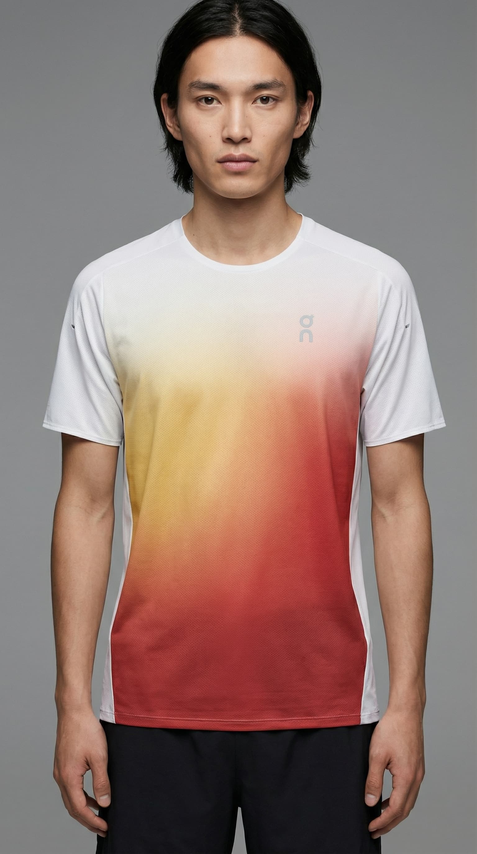 The image is a clean, minimalist fashion portrait with a strong product-first focus.
A model stands in a relaxed, neutral pose, framed from mid-torso up against a smooth gray studio background. The composition is simple and uncluttered, directing full attention to the garment rather than action or environment.
The T-shirt’s gradient print—shifting from white into warm yellow and red—becomes the visual anchor. Its soft fade feels controlled and graphic, almost like a color field, giving the piece a modern, design-led presence. Branding is subtle and restrained, reinforcing a premium, understated tone.
Lighting is even and soft, with minimal shadowing. This keeps fabric texture, fit, and color clarity accurate and readable, typical of high-end lookbook or campaign imagery.
Overall, the image feels modern, calm, and design-forward, sitting at the intersection of performancewear and contemporary fashion. It prioritizes clarity, material quality, and visual balance over storytelling or lifestyle context.