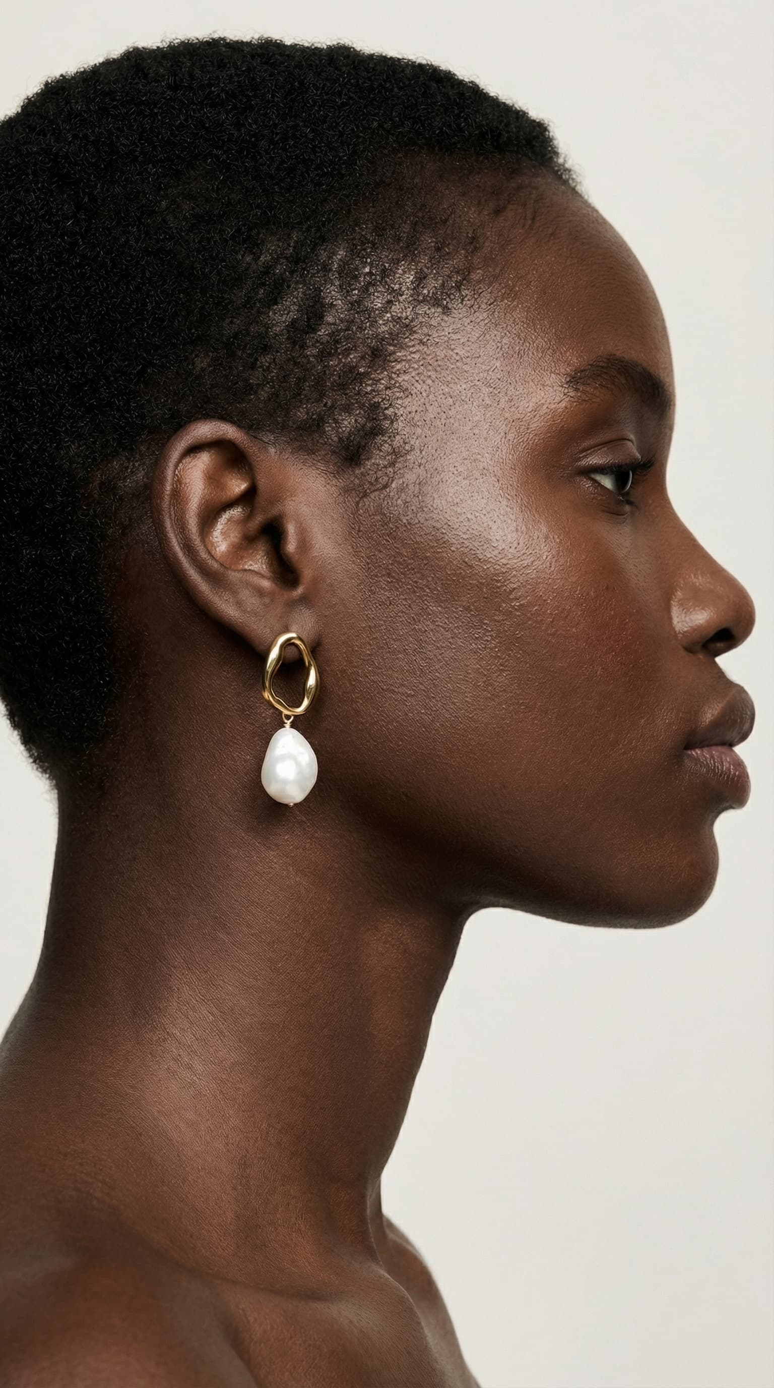 The image is a refined, sculptural beauty portrait focused on form, texture, and restraint.
A close side profile captures the head and neck, cropped tightly so the face is only partially visible. Attention is drawn to the ear, where a circular, geometric earring becomes the focal point. The jewelry’s delicate structure contrasts with the smooth contours of the ear and jawline, reading almost like a small architectural object.
The subject’s hair is cut very short, exposing the natural shape of the head and emphasizing skin and silhouette. Skin texture is softly rendered and luminous, with subtle highlights that define the curve of the neck and cheek without harsh shadow.
The background is a clean, pale neutral, allowing the dark skin tone and silver jewelry to stand out with clarity and elegance. Lighting is controlled and minimal, reinforcing a calm, editorial mood.
Overall, the image feels quiet, precise, and modern, blending fashion and art. It prioritizes shape, material, and negative space over expression or narrative, typical of high-end jewelry or beauty editorials.