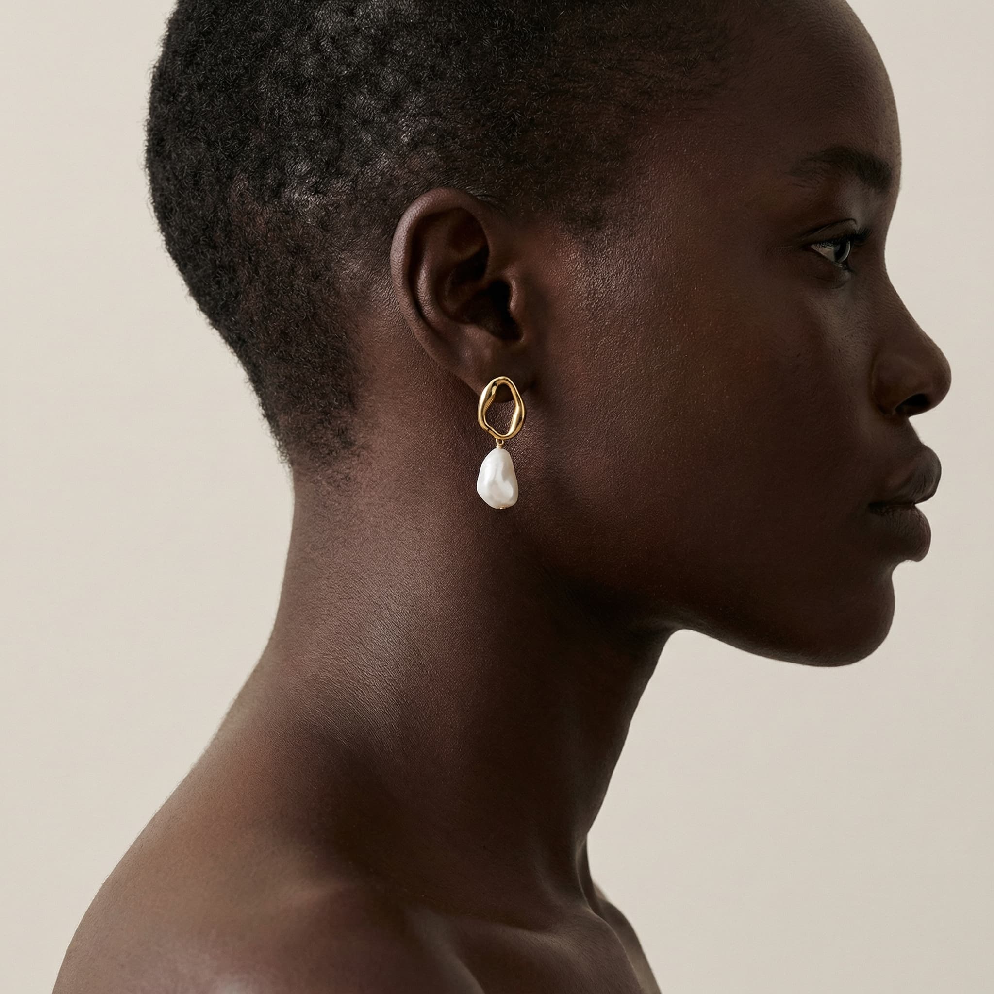 The image is a refined, sculptural beauty portrait focused on form, texture, and restraint.
A close side profile captures the head and neck, cropped tightly so the face is only partially visible. Attention is drawn to the ear, where a circular, geometric earring becomes the focal point. The jewelry’s delicate structure contrasts with the smooth contours of the ear and jawline, reading almost like a small architectural object.
The subject’s hair is cut very short, exposing the natural shape of the head and emphasizing skin and silhouette. Skin texture is softly rendered and luminous, with subtle highlights that define the curve of the neck and cheek without harsh shadow.
The background is a clean, pale neutral, allowing the dark skin tone and silver jewelry to stand out with clarity and elegance. Lighting is controlled and minimal, reinforcing a calm, editorial mood.
Overall, the image feels quiet, precise, and modern, blending fashion and art. It prioritizes shape, material, and negative space over expression or narrative, typical of high-end jewelry or beauty editorials.