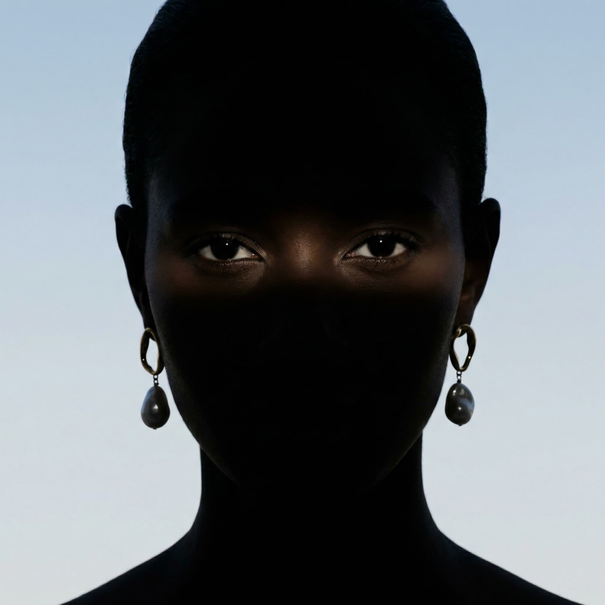 The image is a stark, conceptual portrait built around contrast, concealment, and gaze.
A figure faces the camera directly, but most of the face and head are enveloped in deep shadow or a dark covering, leaving only a narrow horizontal band of light across the eyes. This strip of illumination becomes the emotional and visual center of the image, drawing immediate attention to the gaze.
Large hoop earrings are faintly visible at the sides, their circular shapes subtly breaking the silhouette and adding a graphic fashion element. The background is a soft, open sky tone, which contrasts sharply with the near-black foreground figure and heightens the dramatic separation between subject and space.
The composition is minimal and frontal, almost confrontational, yet restrained. By obscuring identity and emphasizing only the eyes, the image shifts from portraiture to symbolism—suggesting themes of anonymity, protection, mystery, or resistance.
Overall, the image feels powerful, enigmatic, and editorial, using light and shadow as narrative tools. It prioritizes concept and mood over realism, creating a strong, memorable visual statement with very few elements.