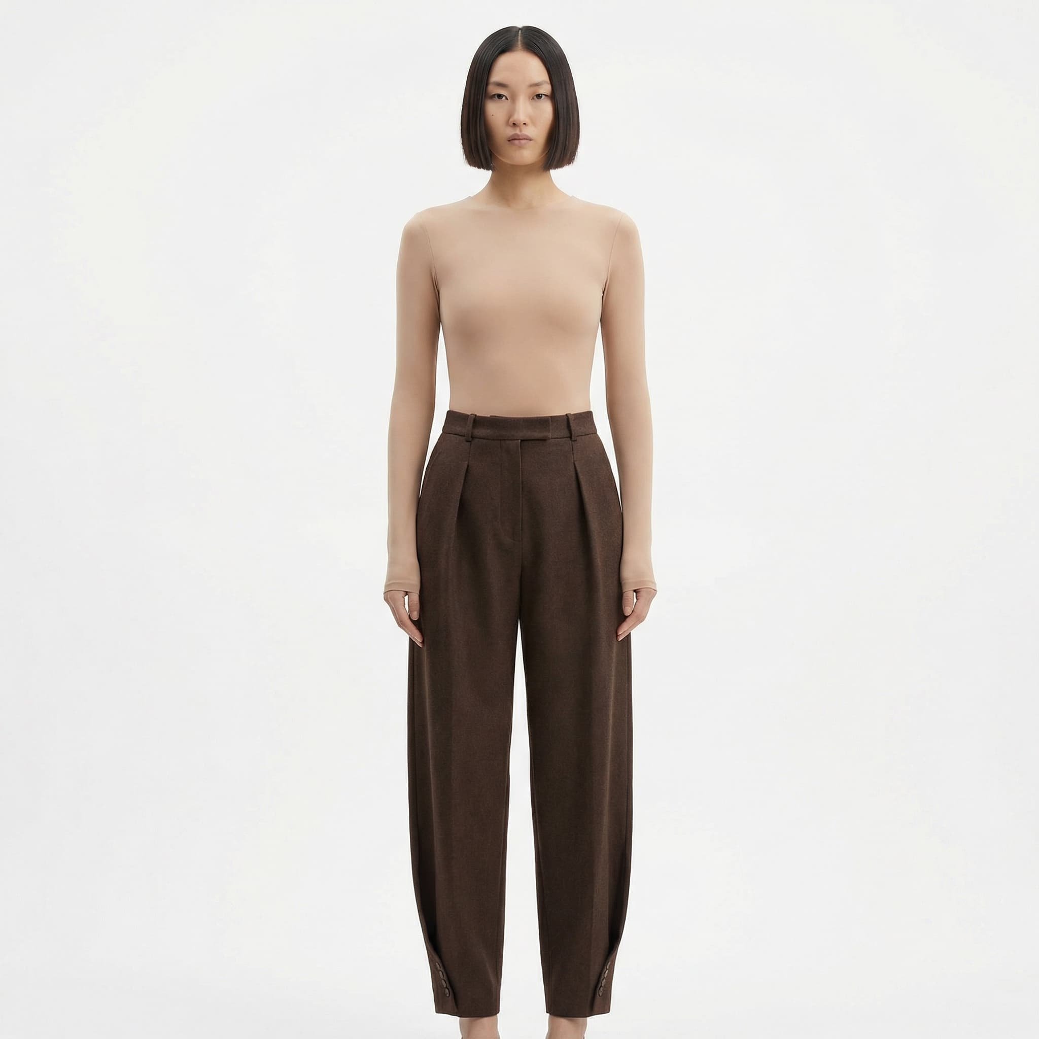 This image is a refined continuation of the minimalist, body-centric studio aesthetic, with a slightly more elevated and composed tone.
The model stands front-facing against a pure white background, centered and symmetrical. The pose is neutral and steady, reinforcing a sense of calm control rather than movement or expression. Eye contact is direct but understated, keeping the mood quiet and self-possessed.
The long-sleeve bodysuit is smooth, matte, and skin-toned, creating a seamless, sculptural silhouette. Its clean neckline and extended sleeves introduce a subtle architectural quality, shifting the focus from bare form to controlled coverage and precision. The garment reads as a second skin—designed to emphasize proportion and line rather than decoration.
Lighting is soft and even, eliminating dramatic shadows and presenting the body and fabric with clarity and honesty. The sharp, blunt haircut adds a graphic counterbalance to the softness of the material, reinforcing the image’s modern, design-led feel.
Overall, the image feels pared-back, intentional, and quietly powerful—typical of contemporary fashion or performancewear lookbooks where neutrality, fit, and form are the message, not narrative or lifestyle.