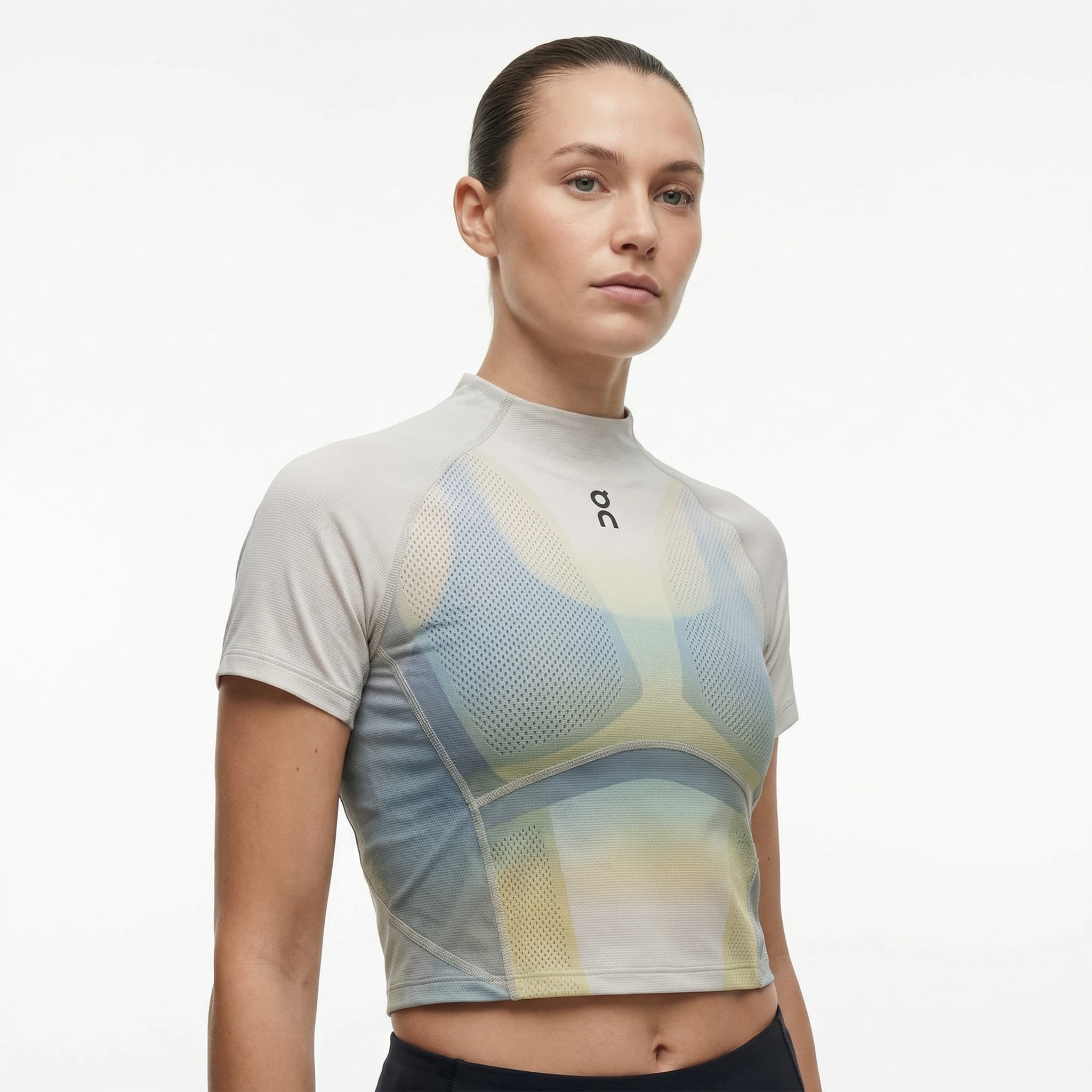 The image is a modern, performance-inspired fashion portrait with a technical, body-aware feel.
A model is shown from the waist up against a clean white background, standing in a relaxed but alert posture. Her gaze is calm and focused, slightly off-camera, reinforcing a composed, athletic presence rather than overt emotion.
The top is the focal point: a cropped, high-neck performance garment with intricate mesh-like patterning and layered color gradients in muted blues, greens, and yellow. The fabric appears engineered rather than decorative, contouring the torso and emphasizing structure, breathability, and movement. Visible seam lines and paneling suggest function and precision.
Styling is minimal—hair pulled back, no accessories—keeping attention on the garment’s construction and the body’s form. Lighting is soft and even, revealing texture and color without harsh contrast.
Overall, the image feels technical, contemporary, and quietly powerful, sitting at the intersection of sportswear and fashion. It communicates control, strength, and design intelligence rather than lifestyle narrative or softness.